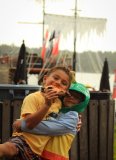 Two smiling kids, one eating pizza, standing by a wooden fence with a ship in the background.